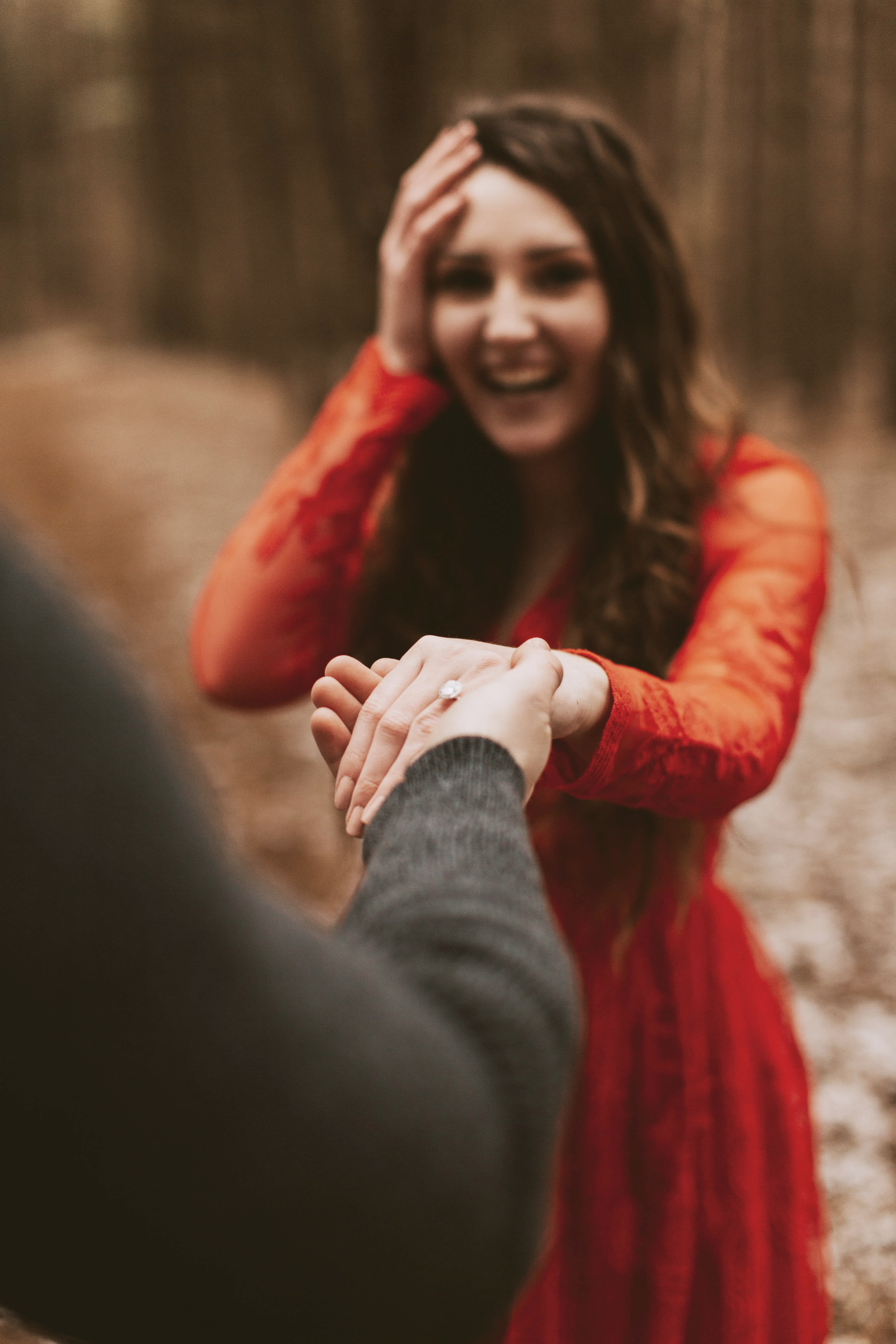 A surprised and happy woman in a red dress extends her hand to receive her new engagement ring from her fiance.