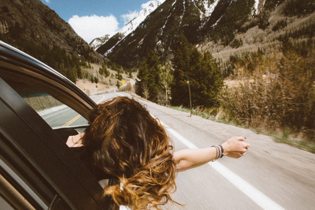 A brunette woman leans her head and arm peacefully out a passenger car window while traveling down a mountain road. 