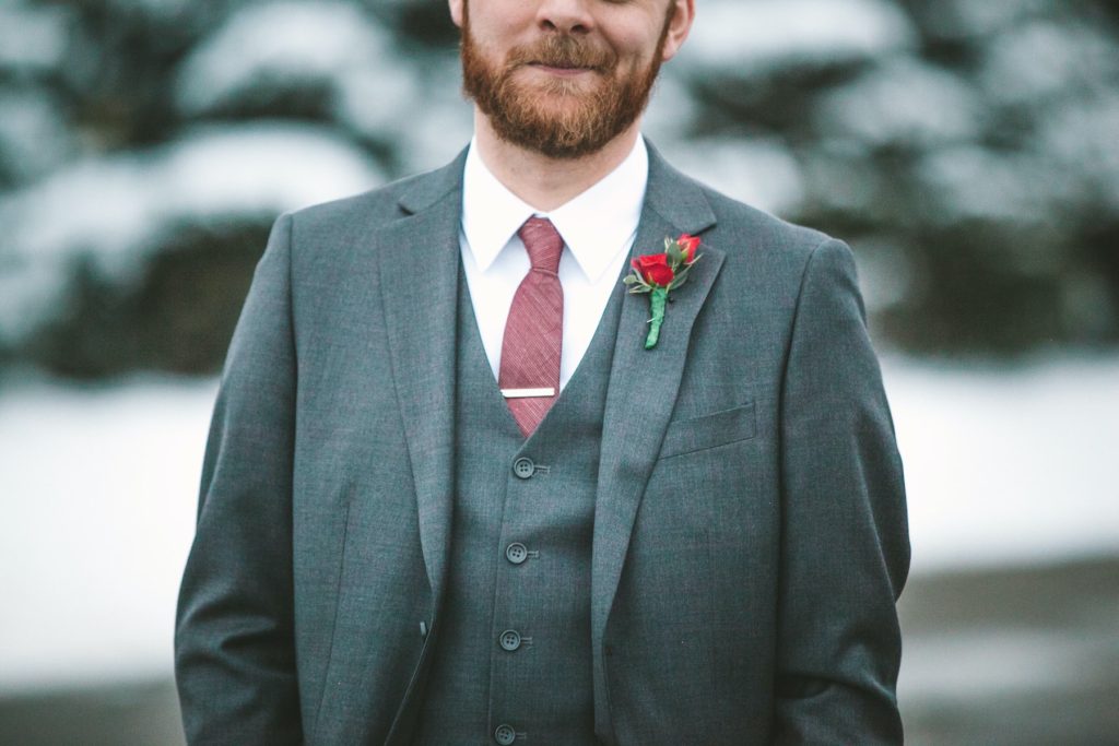 A handsome groom in a gray suit with red accents in front of snow covered fir trees.