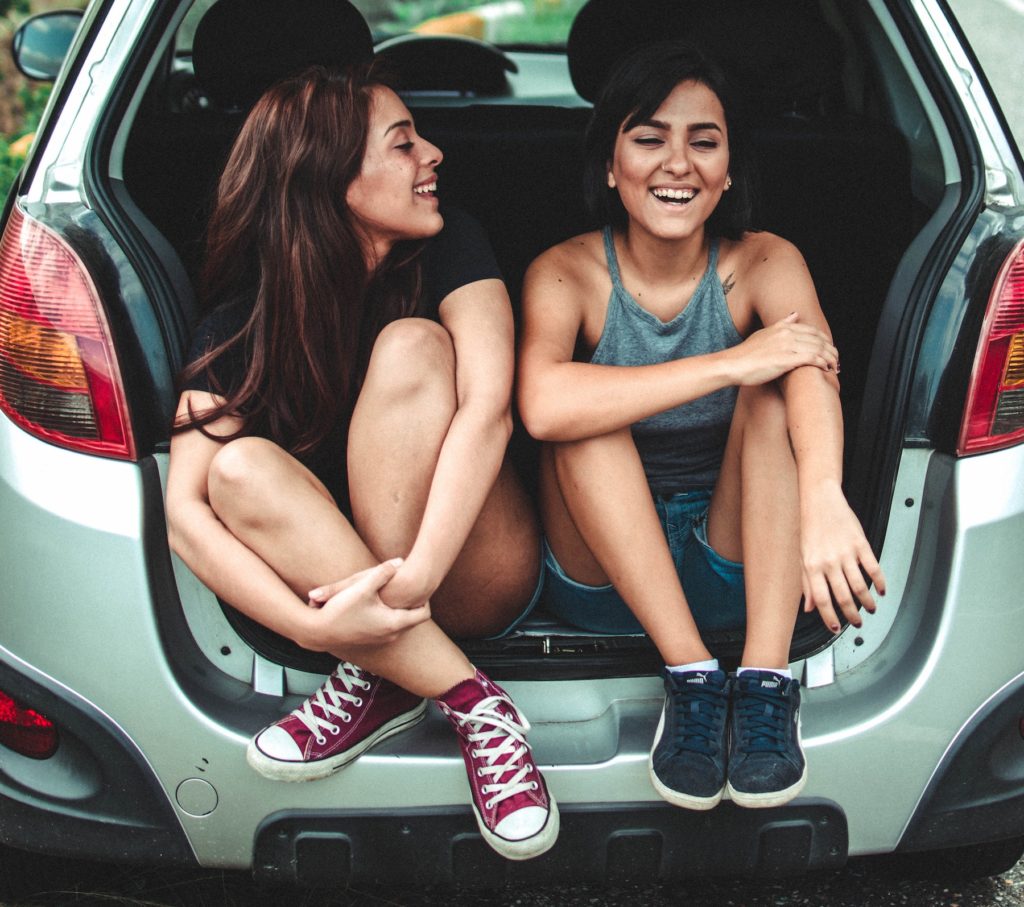 Two smiling women in tennis shoes sitting in the open hatchback of a car.