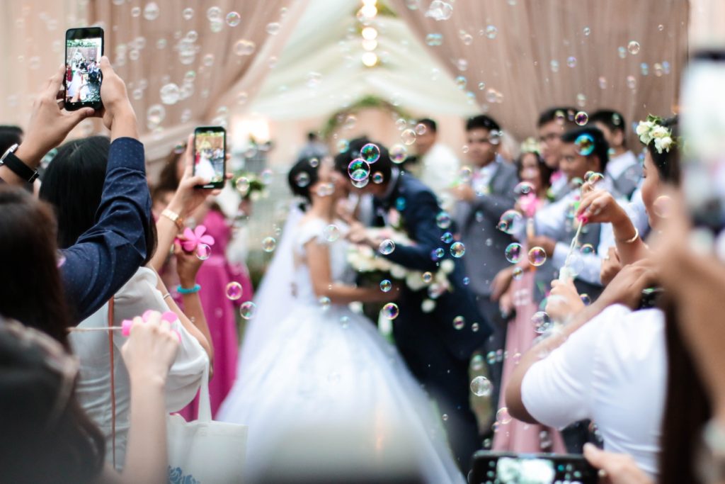 A bride and groom kiss at the end of their wedding ceremony while guests release bubbles and take photos with their cell phones.
