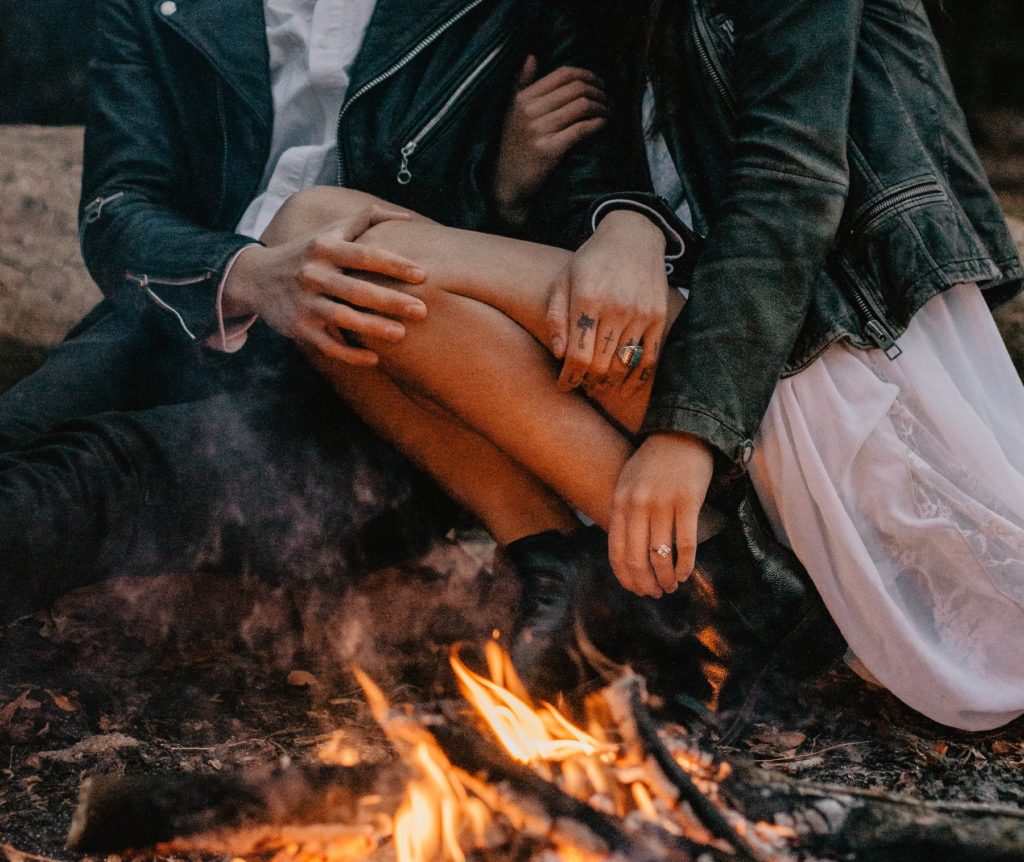 Newlyweds in formal clothes and leather jackets sit by a fire displaying their wedding rings after eloping.