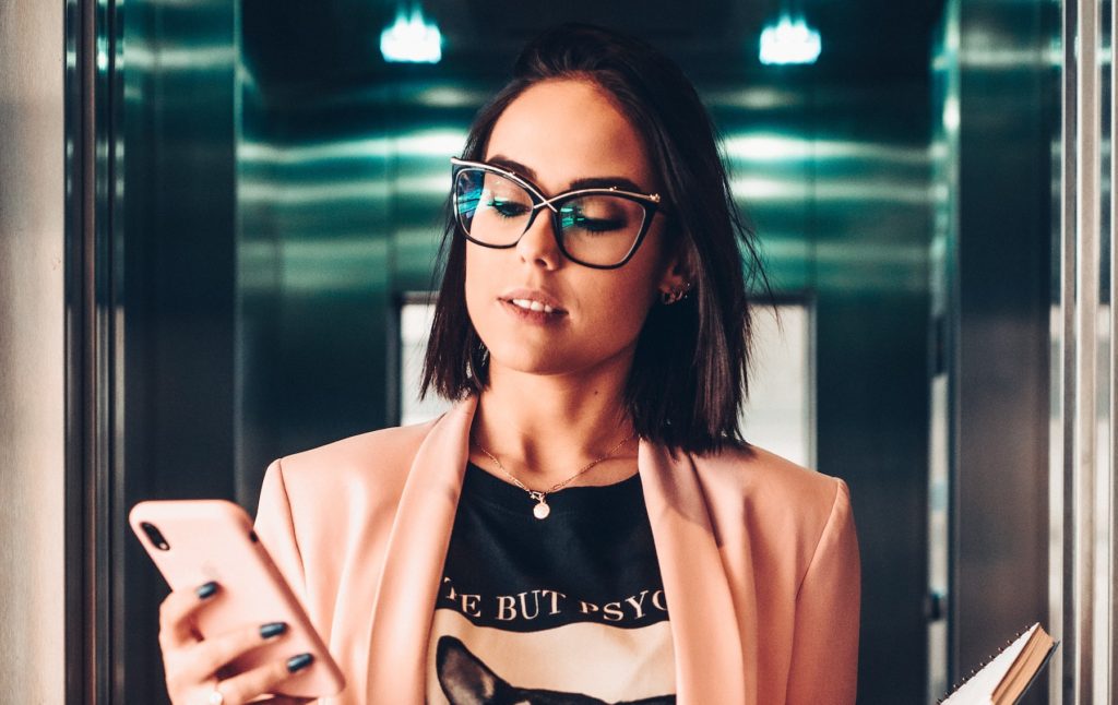 Beautiful brunette woman wearing glasses, T shirt, and pink blazer observes her phone on the way to work.