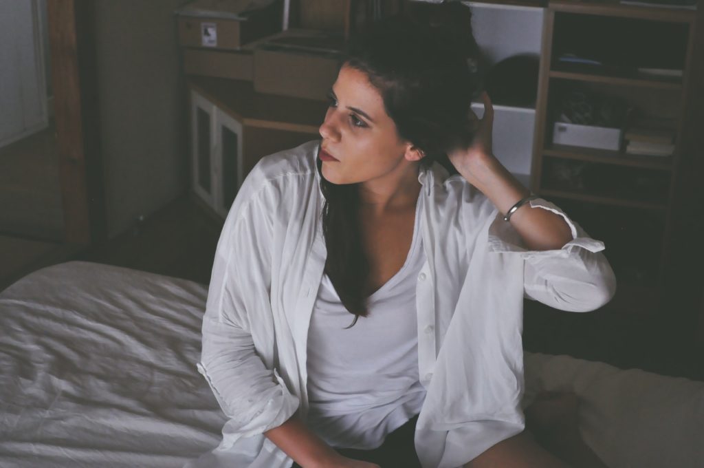 A woman in white pushes her hair back off her shoulder while sitting in her apartment.