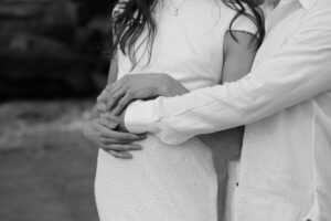 Young newlywed couple, faces cropped, man in dress shirt and woman in bridal dress cradling baby bump