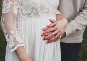 Pregnant woman with her hand overtop her partner's on her baby bump, dressed in white with wedding rings visible