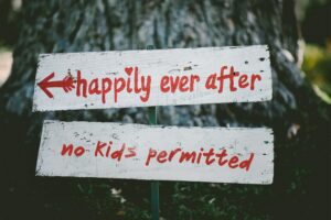 A white painted wedding sign in front of a tree reading in red paint "happily ever after" and then "no kids permitted" underneath.