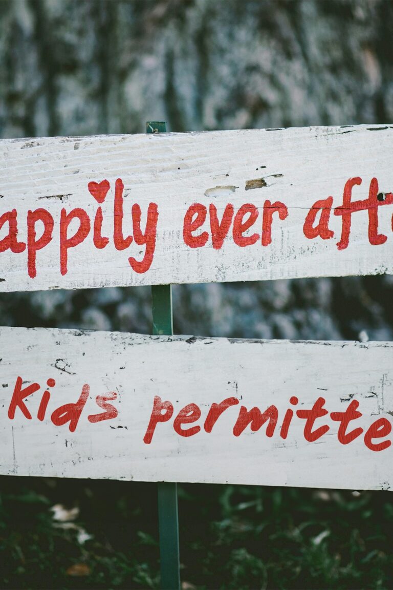 A white painted wedding sign in front of a tree reading in red paint "happily ever after" and then "no kids permitted" underneath.