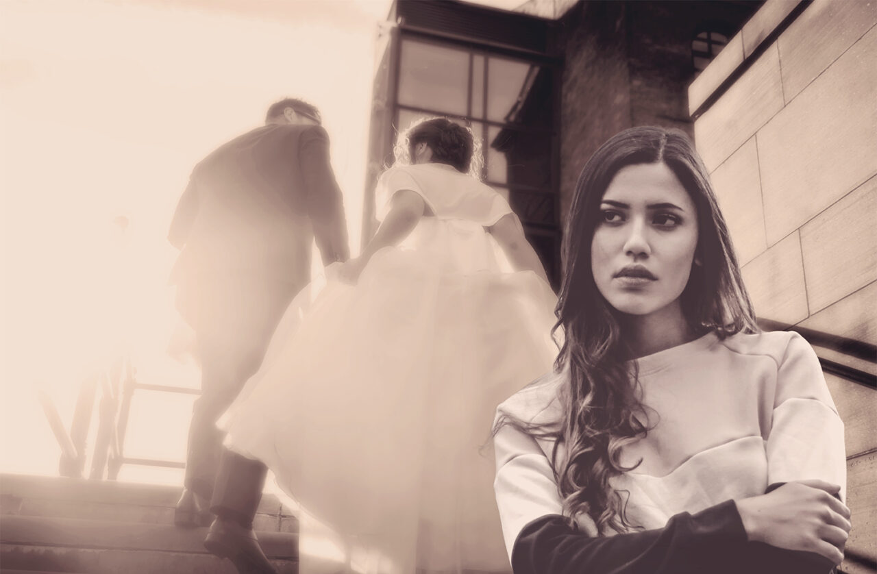 young brunette woman looking annoyed with arms folded while a bride and groom elope behind her on a staircase