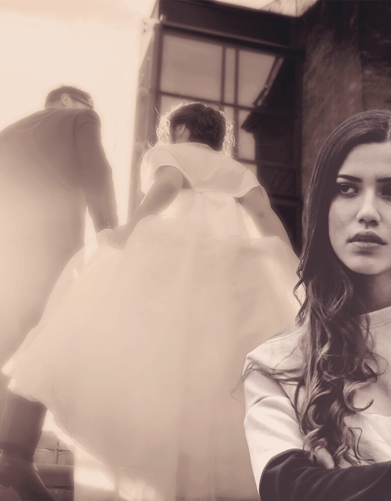 young brunette woman looking annoyed with arms folded while a bride and groom elope behind her on a staircase