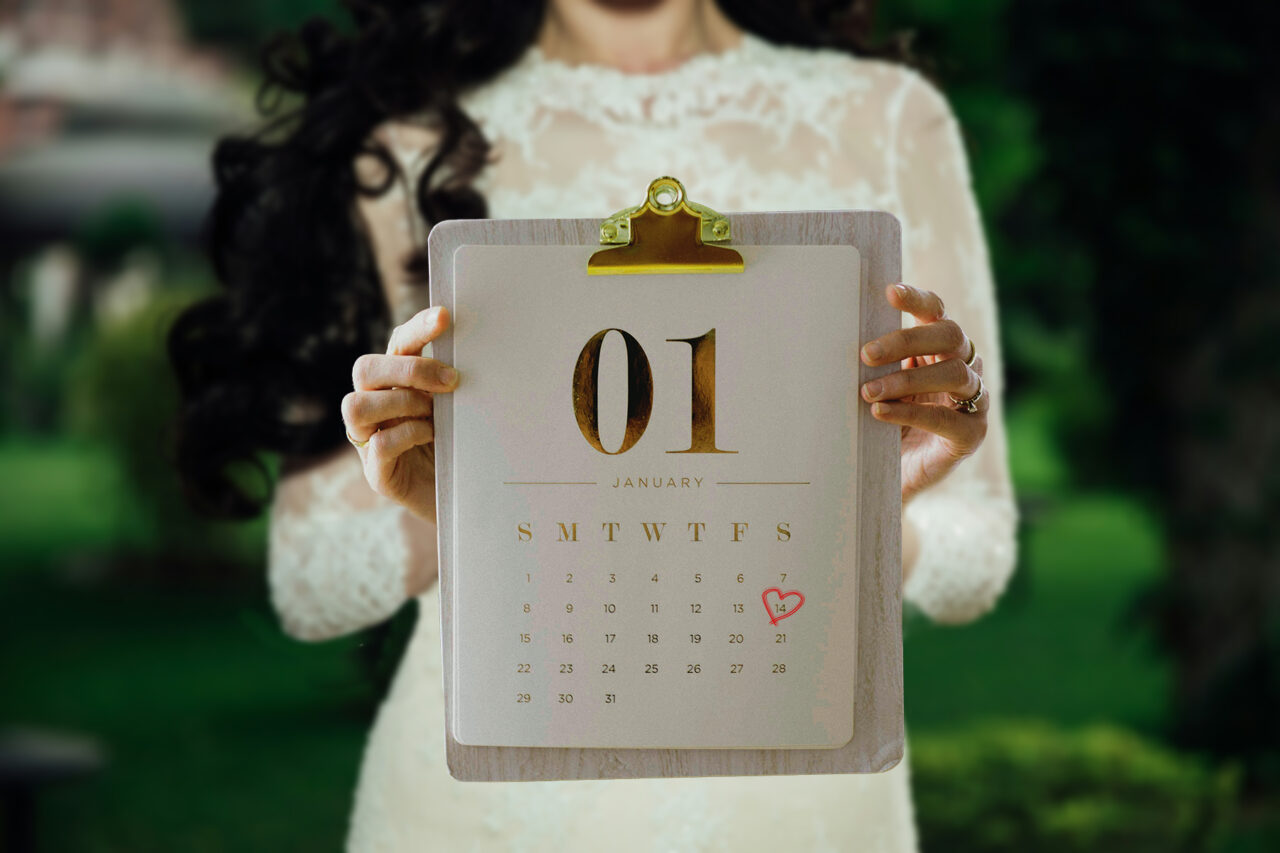 bride wearing a wedding gown in background, holding a calendar on a clipboard in foreground with her wedding date circled in a red heart