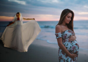 pregnant bridesmaid on a beach while bride poses in gown behind her during sunset