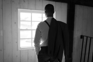 black and white moody portrait of a groom standing in front of a window with suit jacket thrown over shoulder