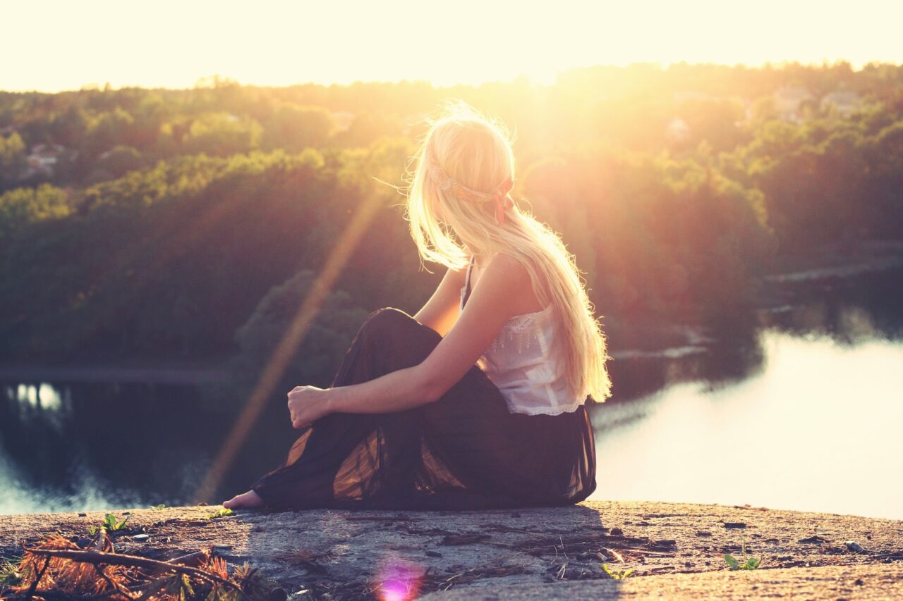 A thoughtful young blonde woman looking out over a river during sunset from atop a cliff.