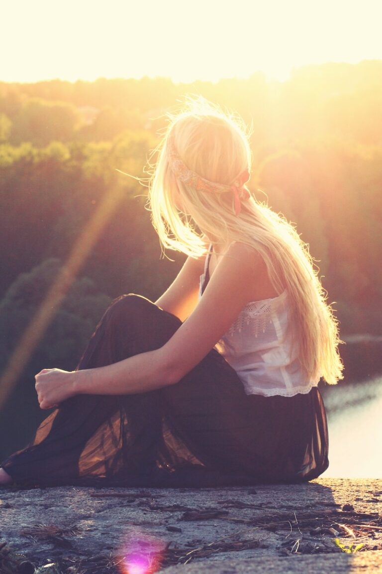 A thoughtful young blonde woman looking out over a river during sunset from atop a cliff.