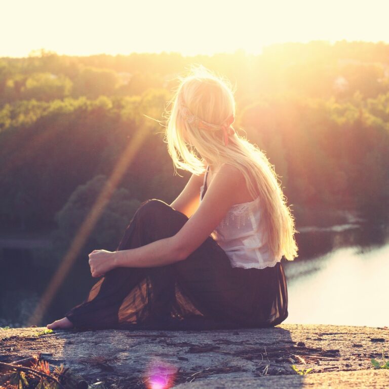 A thoughtful young blonde woman looking out over a river during sunset from atop a cliff.